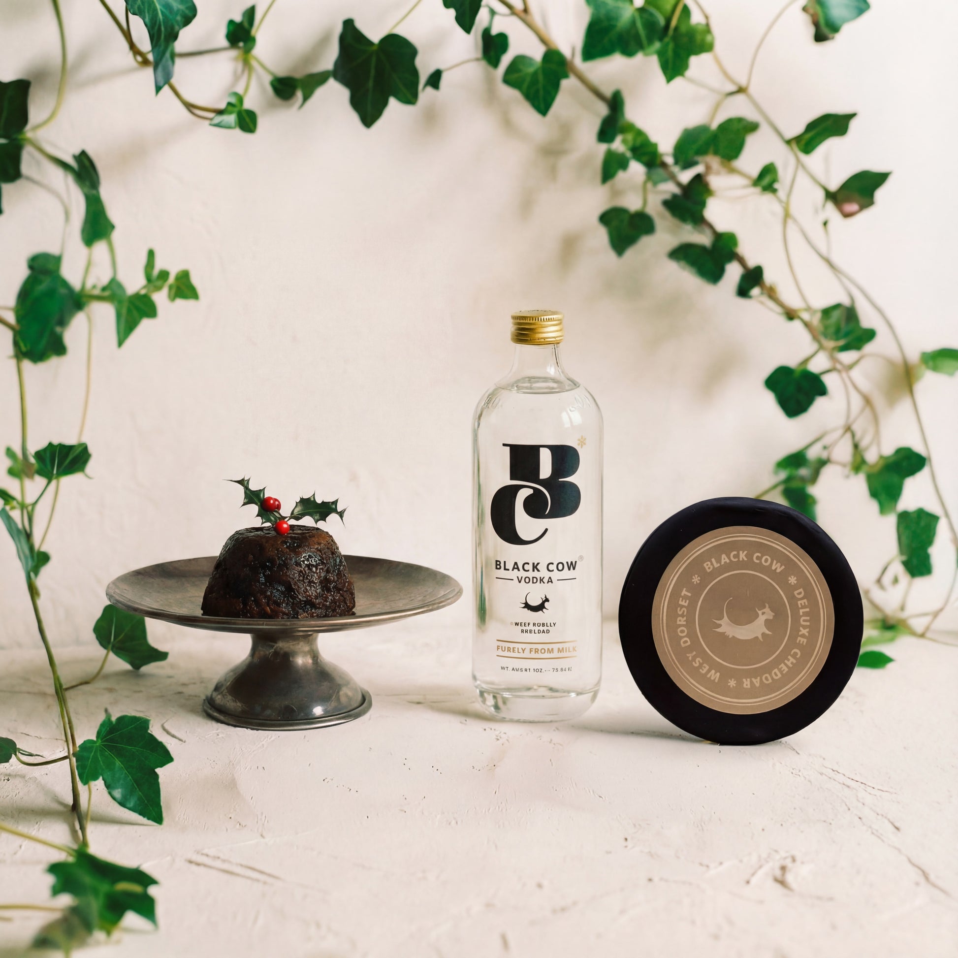 Bottle of Black Cow gin with a Christmas pudding and a coaster on a white surface with ivy in the background.