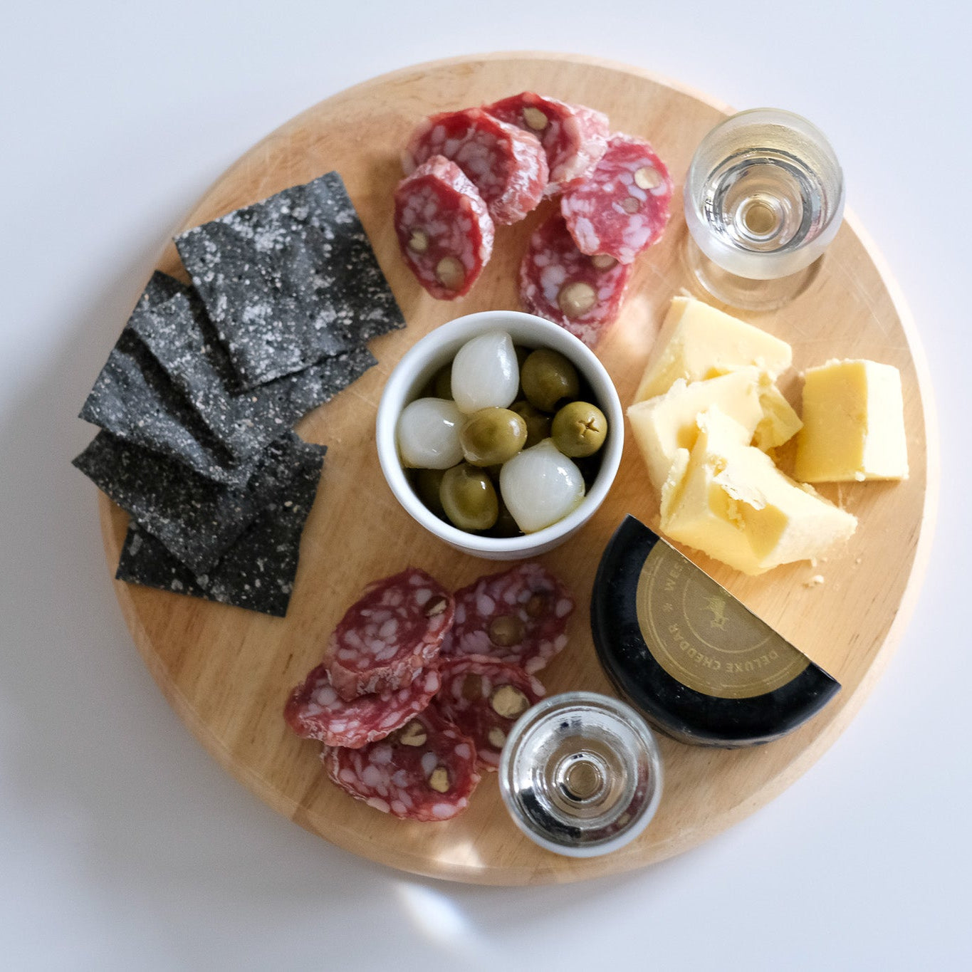 Wooden charcuterie board with various foods including salami, cheese, and olives on a white background.