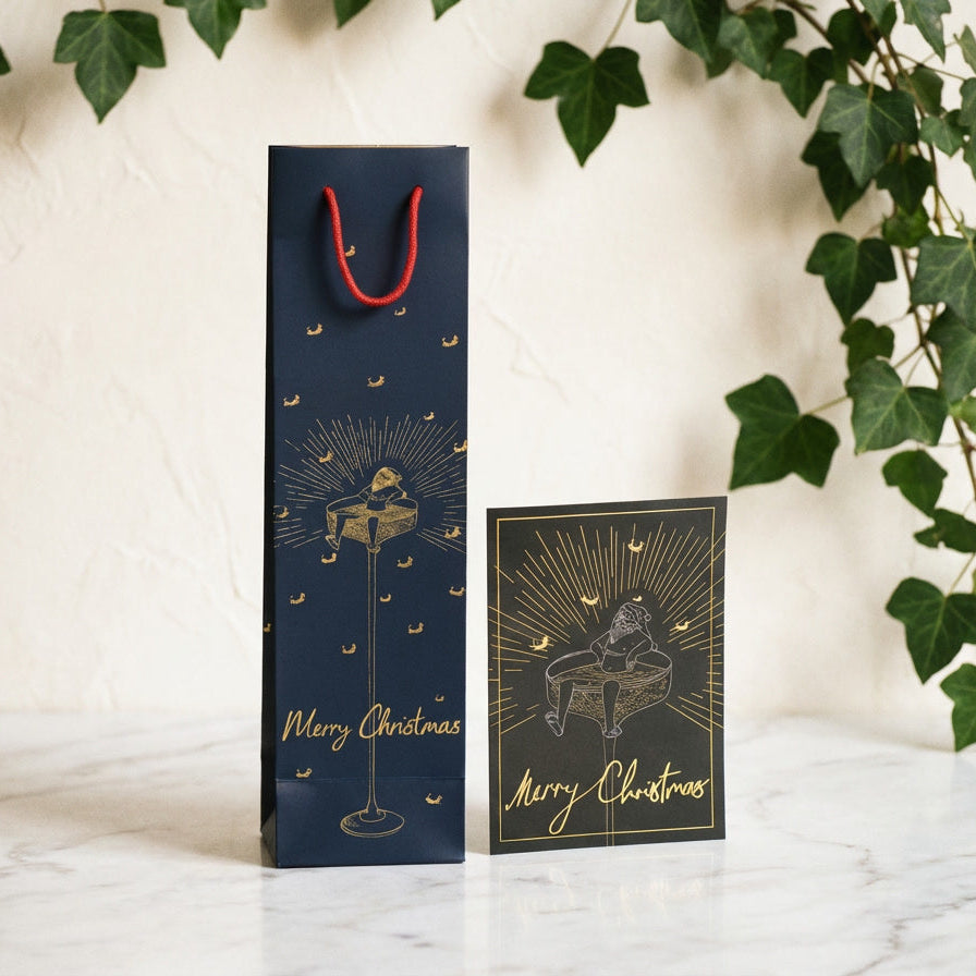Navy blue gift bag with gold festive design and 'Merry Christmas' text, accompanied by a matching card, on a light background with greenery.